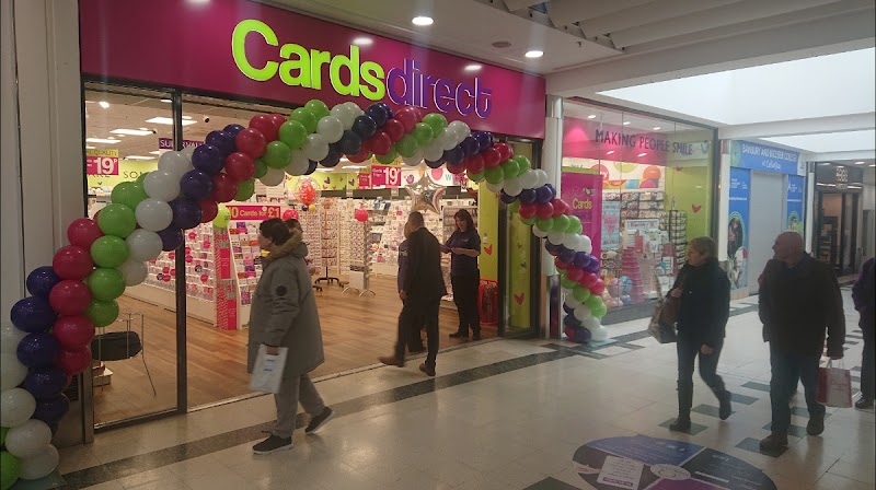 People walk past a Cards Direct store decorated with a balloon arch inside a shopping mall.